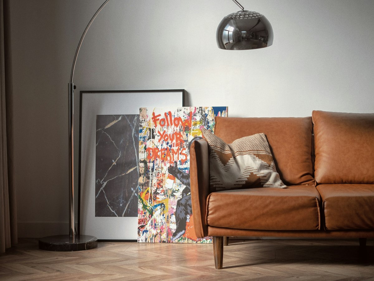 A large arc floor lamp with a chrome shade beside a tan leather sofa, with framed art leaning against the wall.